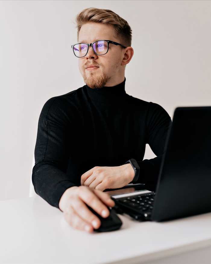 A blond man in glasses and a black turtleneck working at a laptop at a white desk