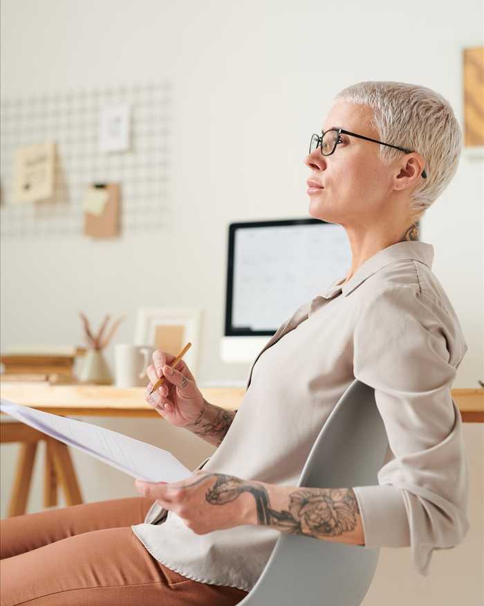 A woman with short silver hair and glasses sitting back in a chair, thinking with papers and a pencil in her tattooed hands