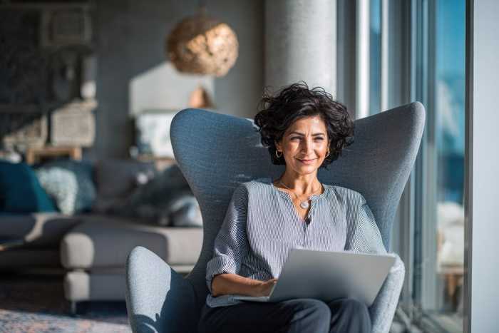 A woman with dark curly hair smiling in a grey wing chair with a laptop