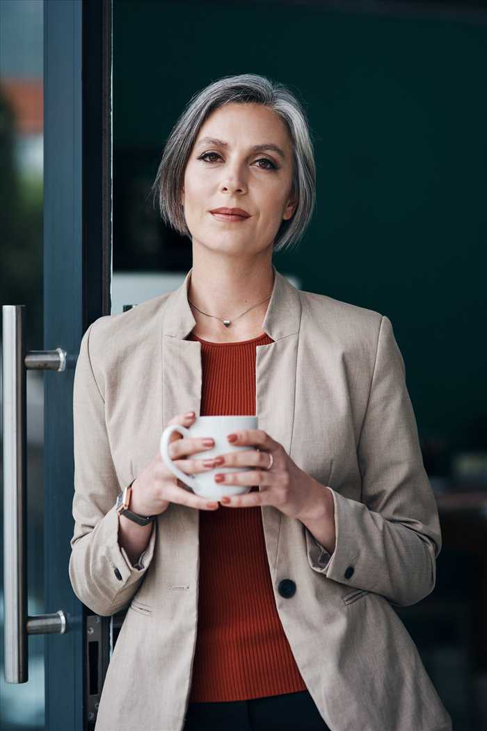 A grey-haired woman in a beige blazer holding a white mug and leaning on a glass door