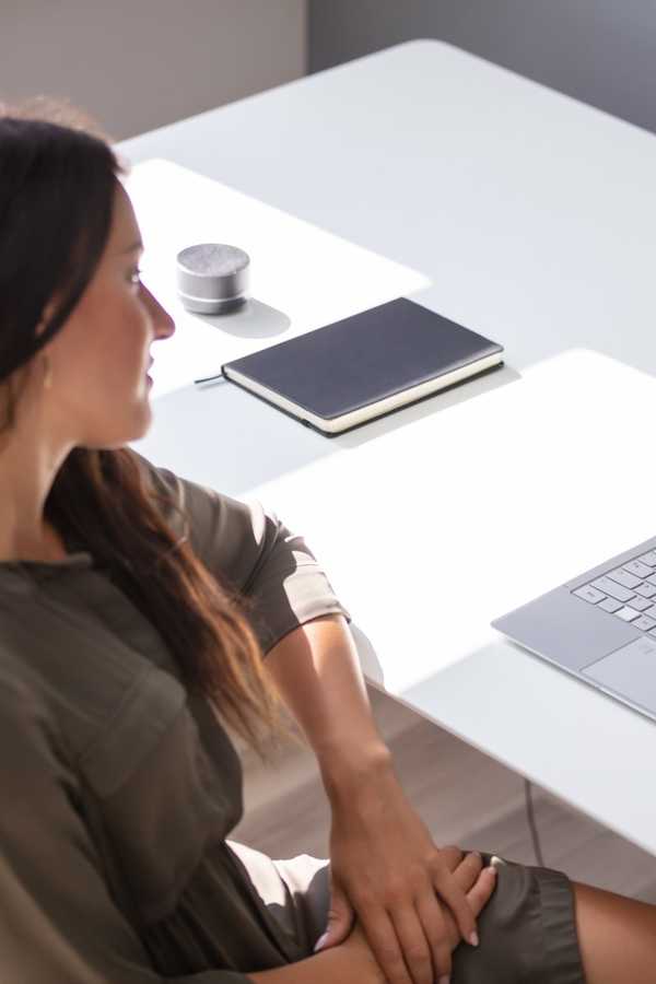A woman seated at a white desk with a notebook and laptop, in soft daylight