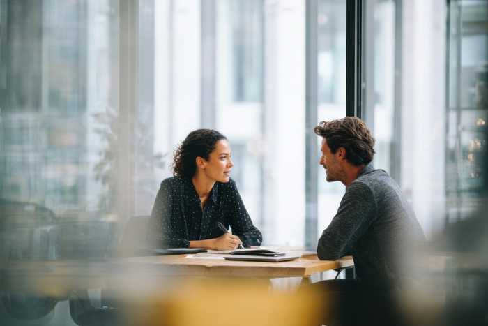 A woman and a man in conversation across a table by a large window
