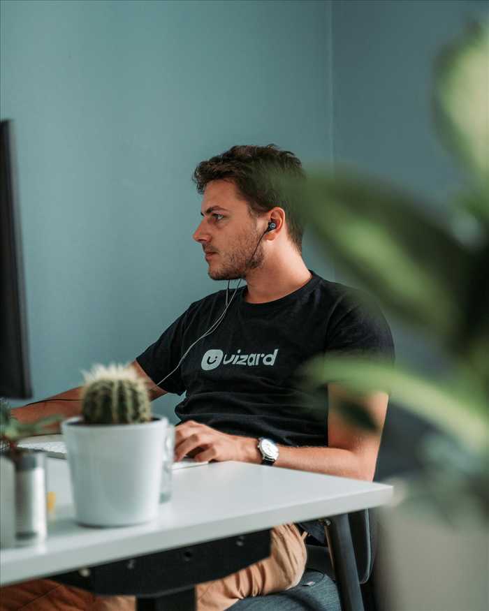 A man in a black t-shirt and earbuds working at a desk by a monitor, with a cactus and plants in the foreground