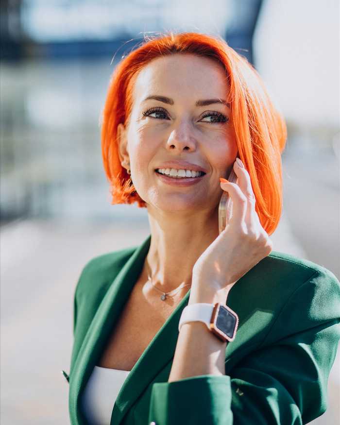 A smiling woman with bright orange hair in a green blazer talking on her phone outdoors