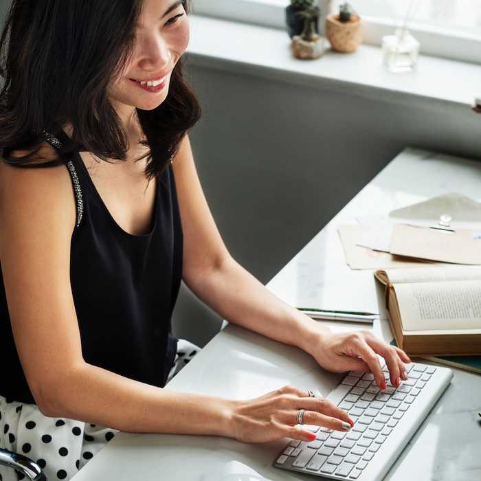 A woman in a black top smiling as she types on a white keyboard at a bright desk