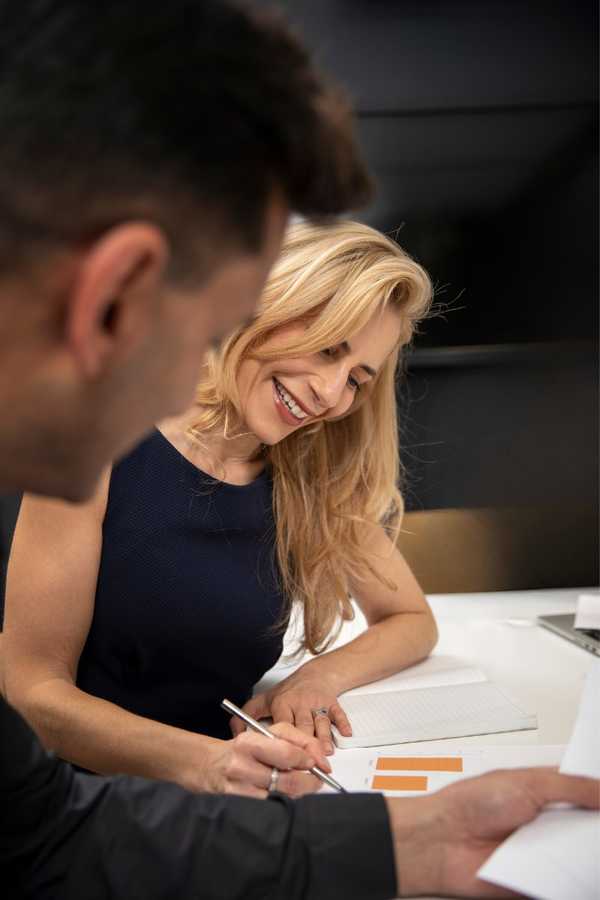 A smiling blonde woman in a navy top writing at a desk beside a colleague reviewing papers